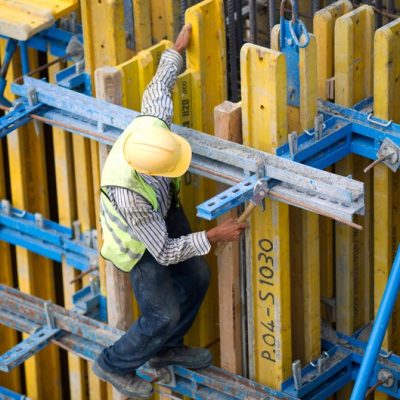 Worker in safety helmet and reflective vest climbing scaffold with yellow and blue pallet racking systems, emphasizing safety and industrial maintenance in warehouse environments.